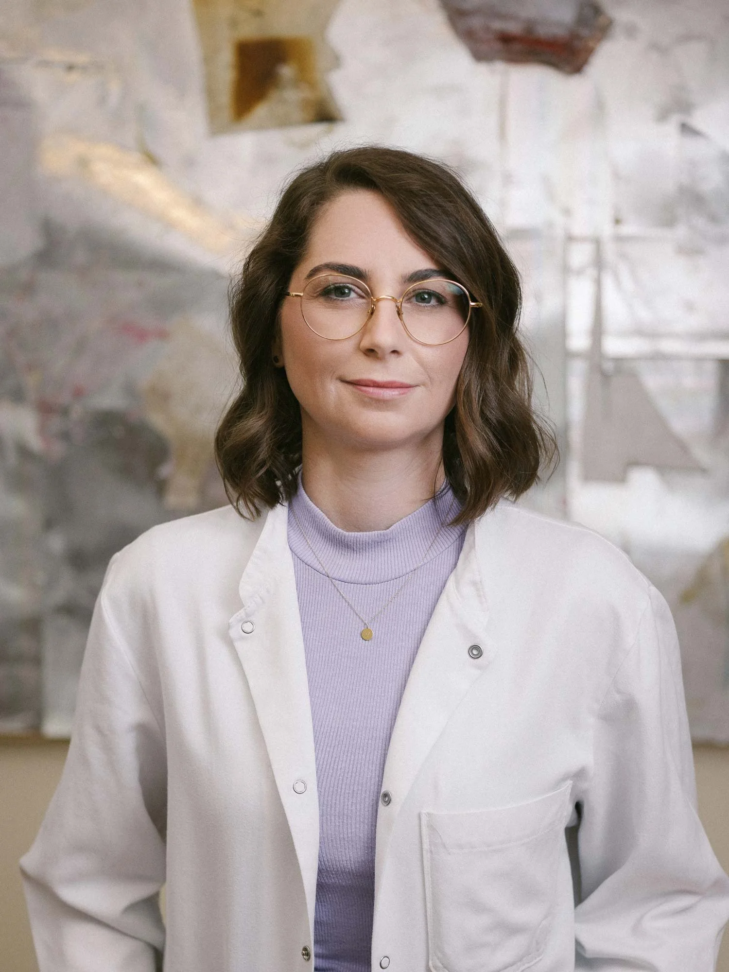 Eine junge Frau mit braunen, lockigen Haaren, Brille und einem weißen Kittel posiert vor einer Wand mit abstrakten Wandbildern.