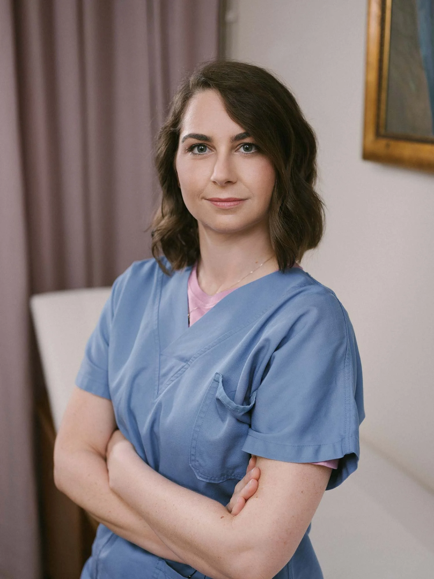 Eine junge Frau in blauer Krankenschwesteruniform steht in einem Krankenzimmer mit verschränkten Armen und schaut in die Kamera.