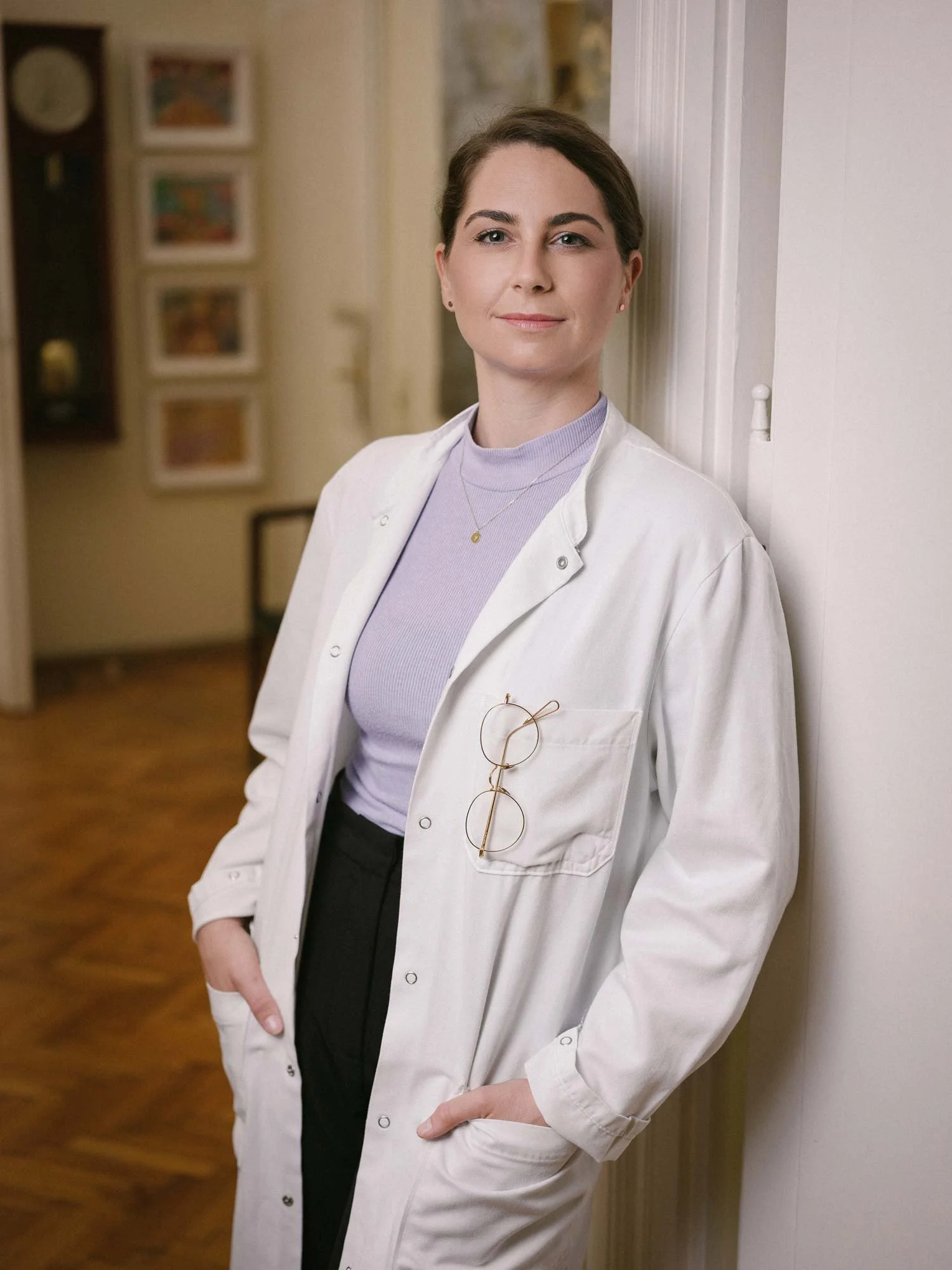 Eine junge Frau trägt einen weißen Laborkittel mit einer Brille in der Brusttasche, steht an einer weißen Wand und lächelt leicht.