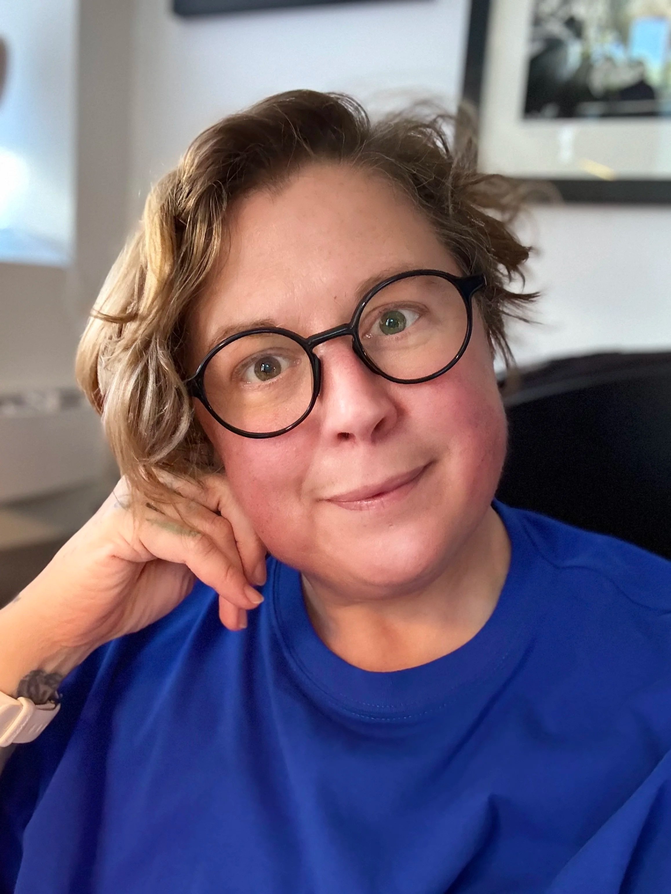 A nonbinary white person in a royal blue shirt and glasses, from the shoulders up. They are resting their cheek on their fist with their head tilted, smiling.