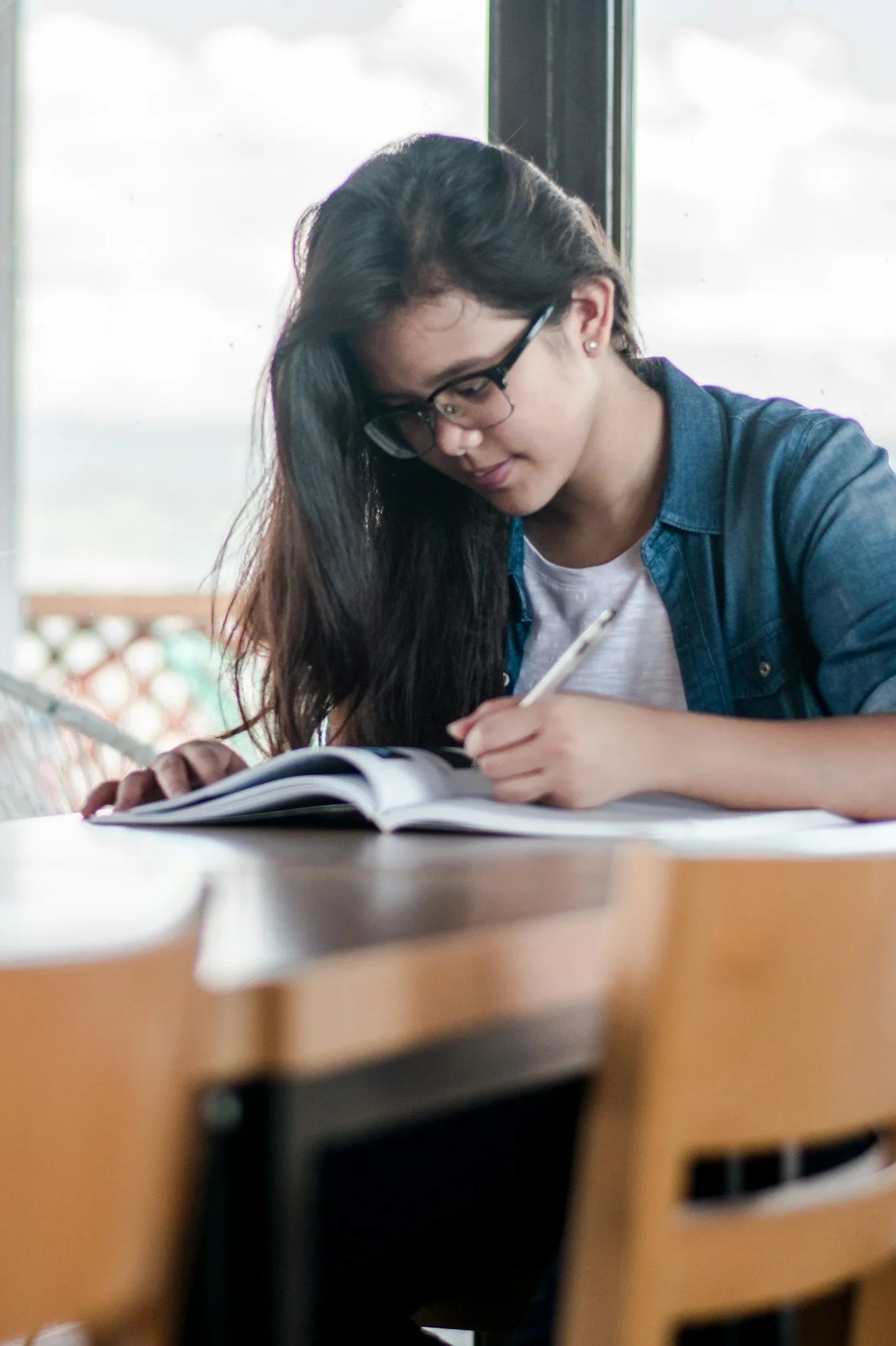 Student wearing glasses writing in an open notebook at a classroom desk