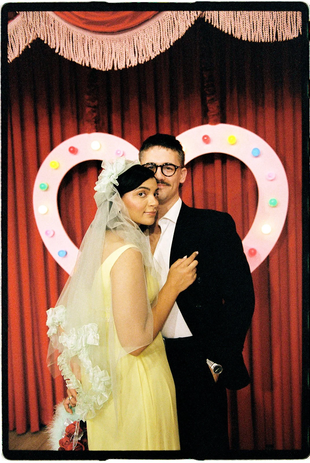 Couple posing together in front of a pink heart-shaped arch and red velvet curtains at Sure Thing NYC.