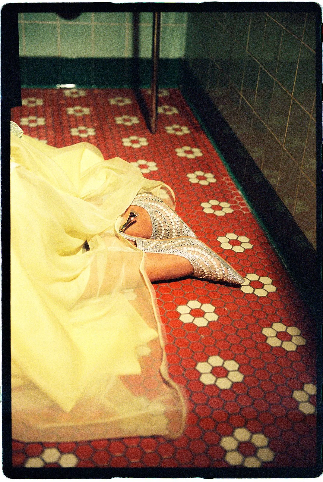 Close-up of sparkling silver bridal heels and a pale yellow wedding dress against retro red and white floral floor tiles.