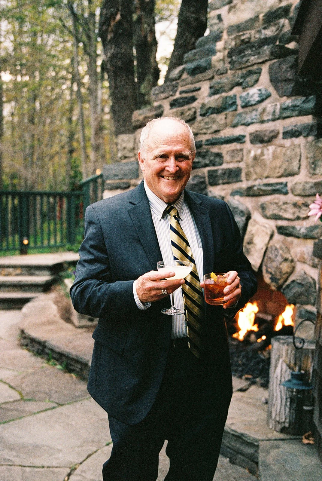 Candid film portrait of a wedding guest holding two cocktails beside the outdoor stone fireplace at Deer Mountain Inn.