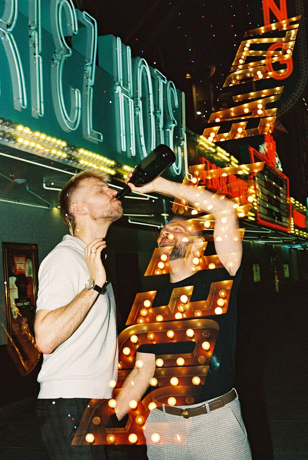 Man giving his husband a drink of champagne straight from the bottle during their Fremont Street elopement photos in Las Vegas on 35mm film.