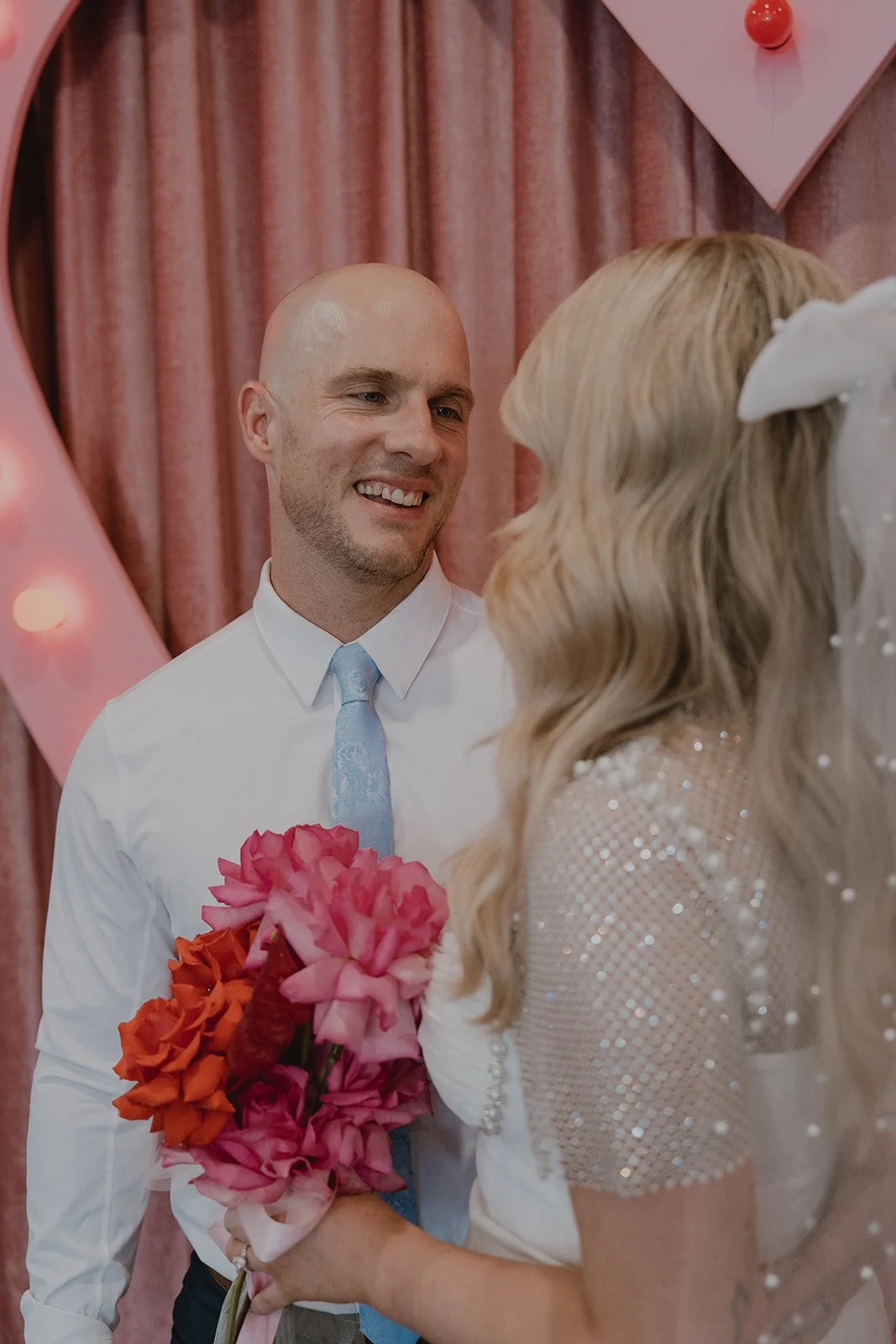 Groom smiling at the bride during portraits inside Sure Thing Chapel with pink velvet curtains behind them.