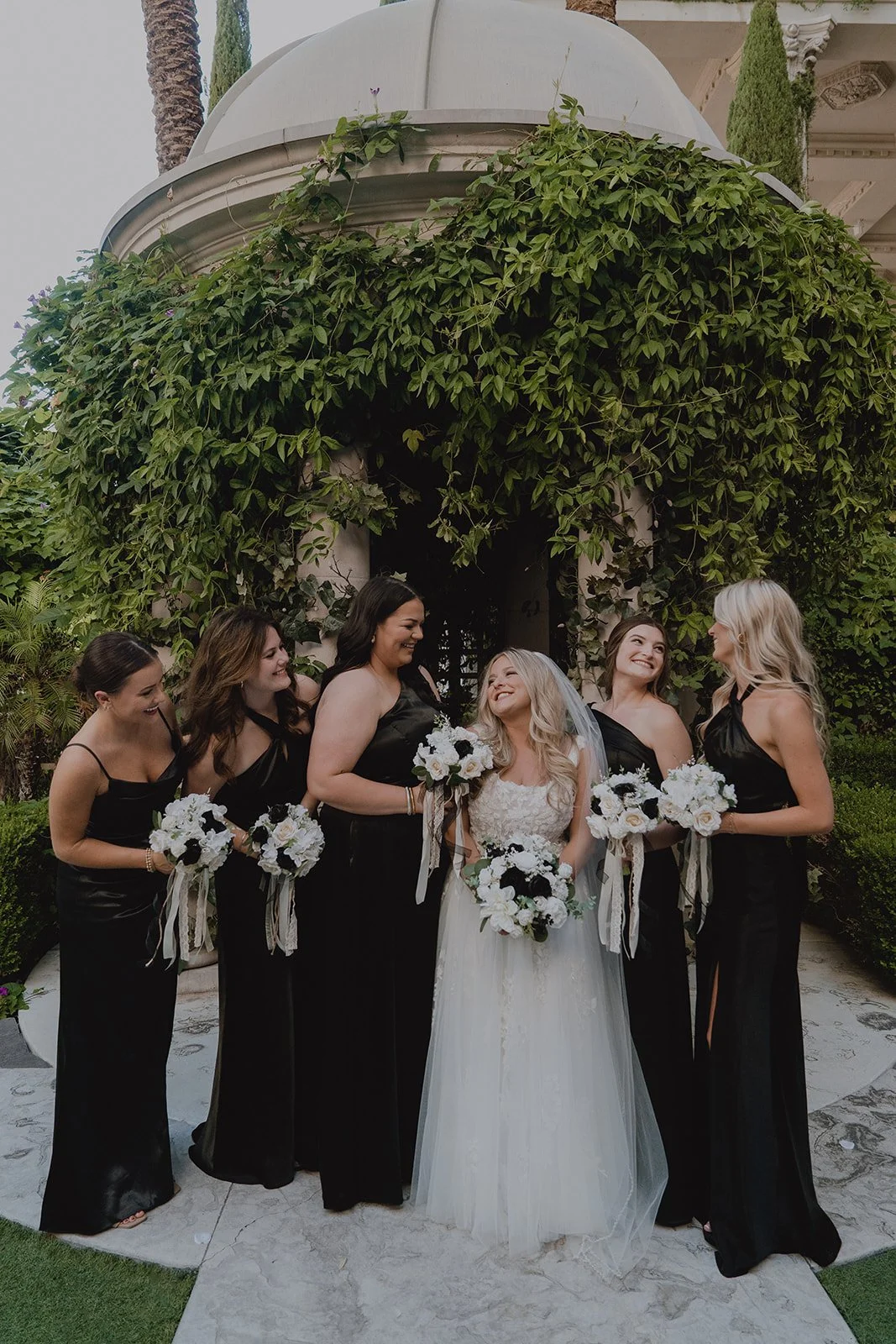 Bridesmaids in sleek black dresses posing with the bride in front of the ivy-covered Venus Garden at Caesars Palace in Las Vegas.