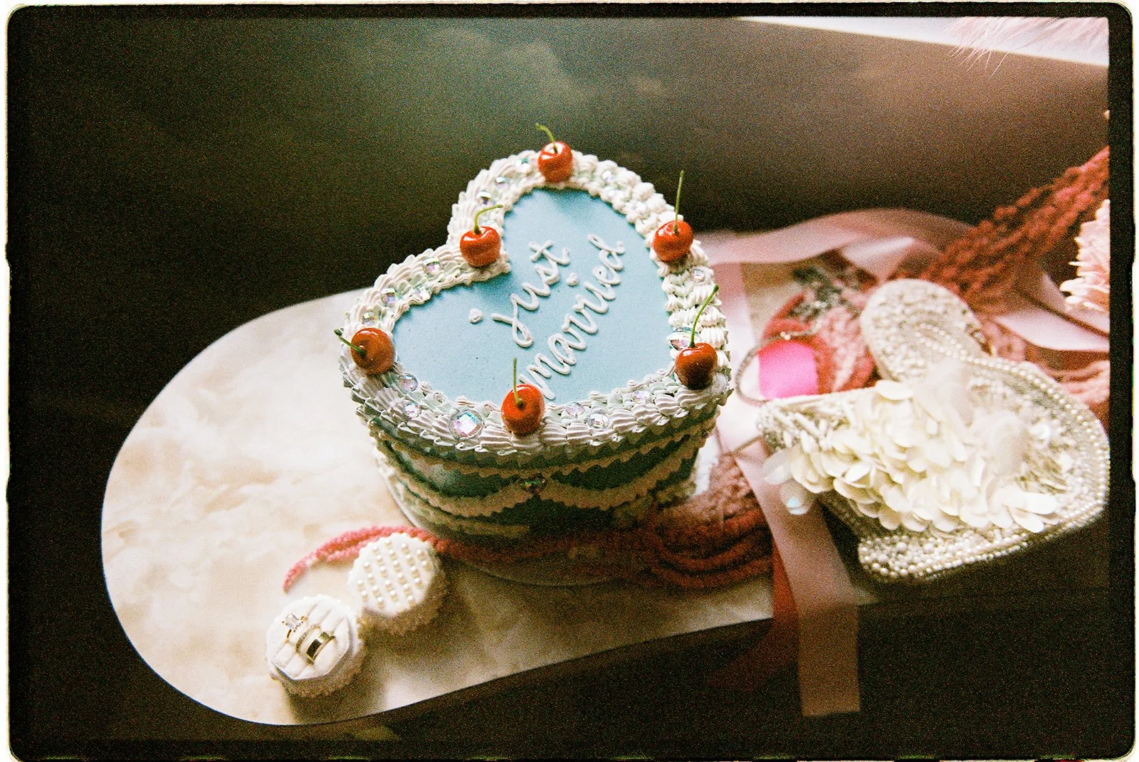 Heart-shaped wedding cake with cherry decorations, surrounded by colorful bridal accessories and flowers beside a chapel window.
