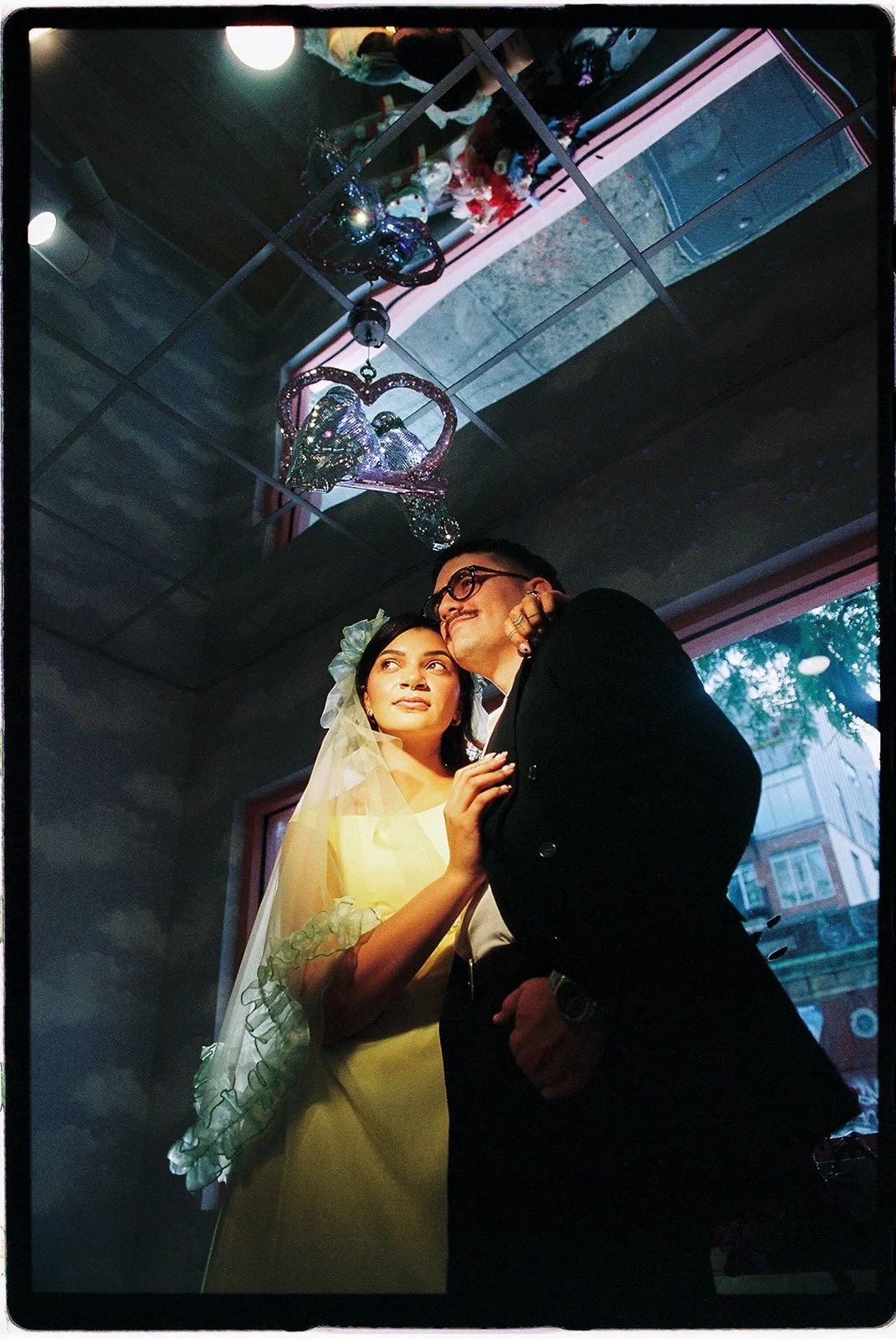 Moody film wedding portrait of a couple embracing beneath a vintage silver bird decoration inside Sure Thing NYC.