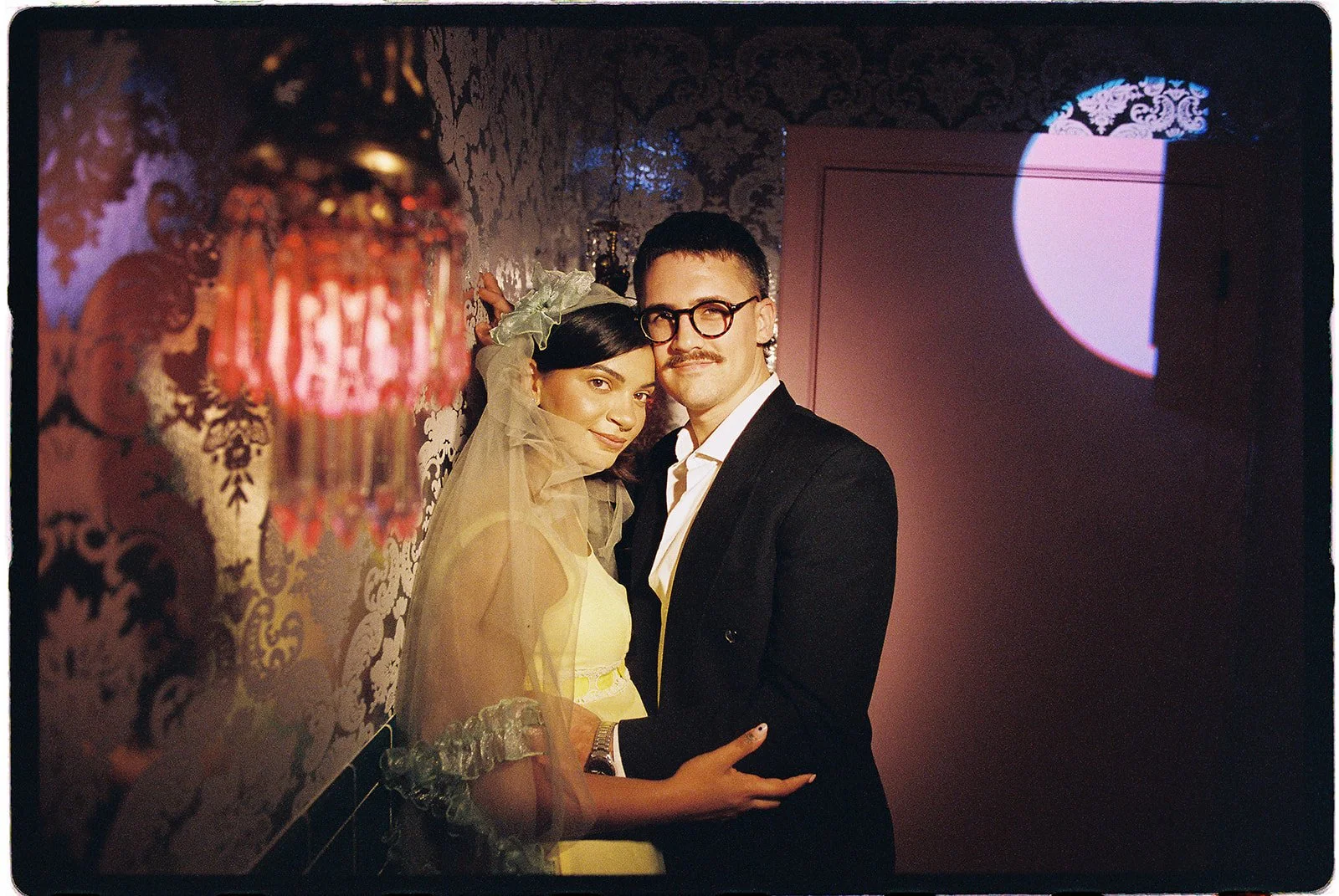 Couple posing together in a dimly lit room with vintage patterned wallpaper and warm red lighting at Sure Thing NYC.