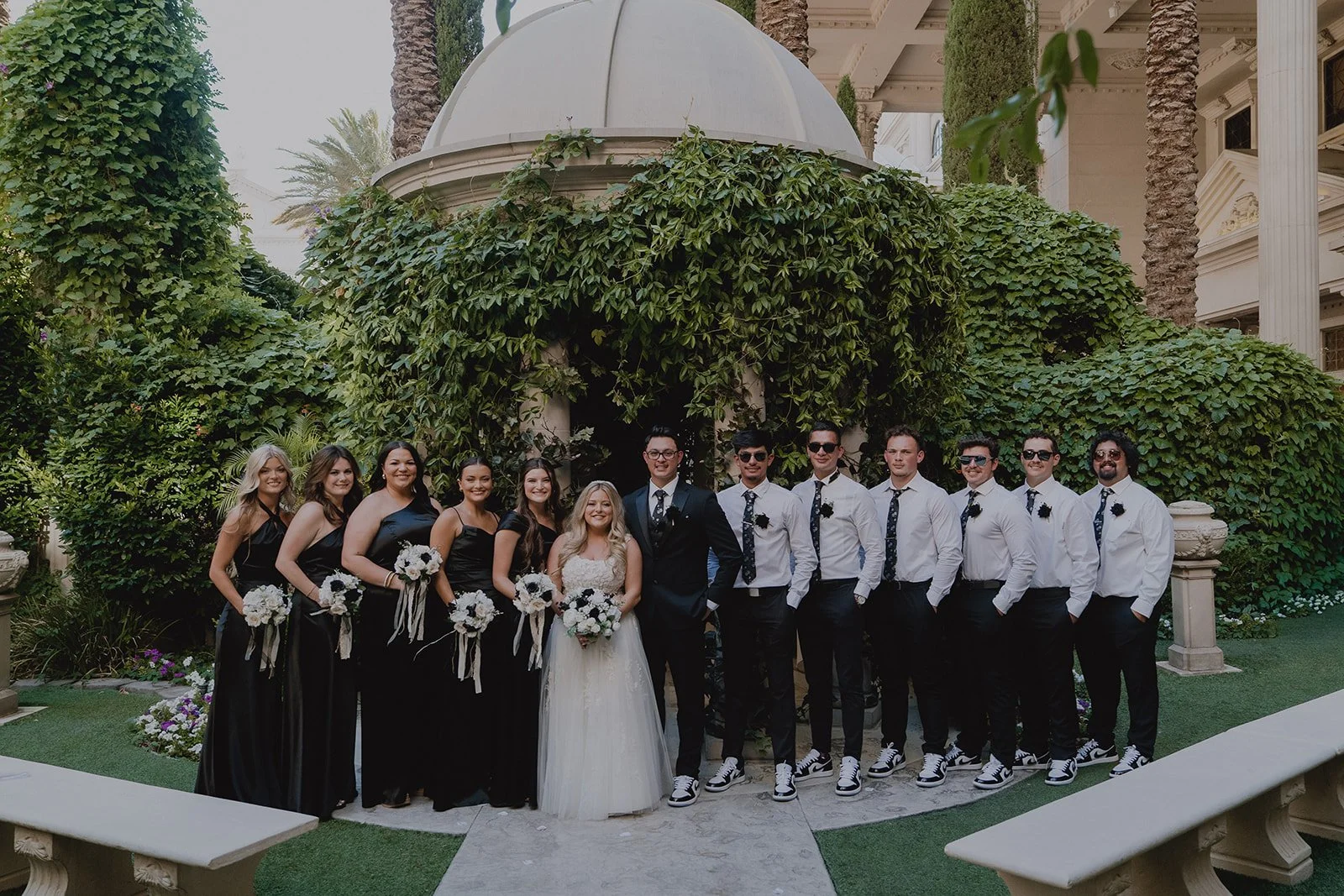 Wedding party group photo at Caesars Palace Venus Garden with bride, groom, bridesmaids in black dresses, and groomsmen in black and white attire surrounded by greenery.