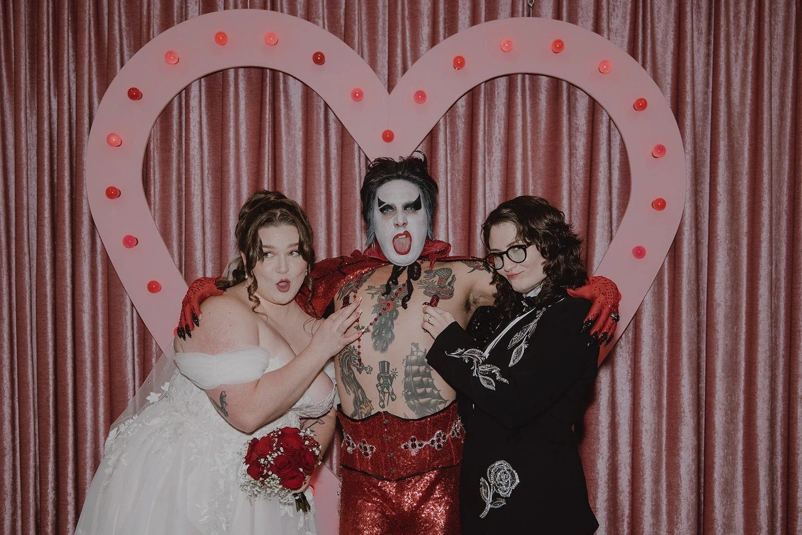 Couple posing with Hellvis in front of the pink heart backdrop at Sure Thing Chapel.