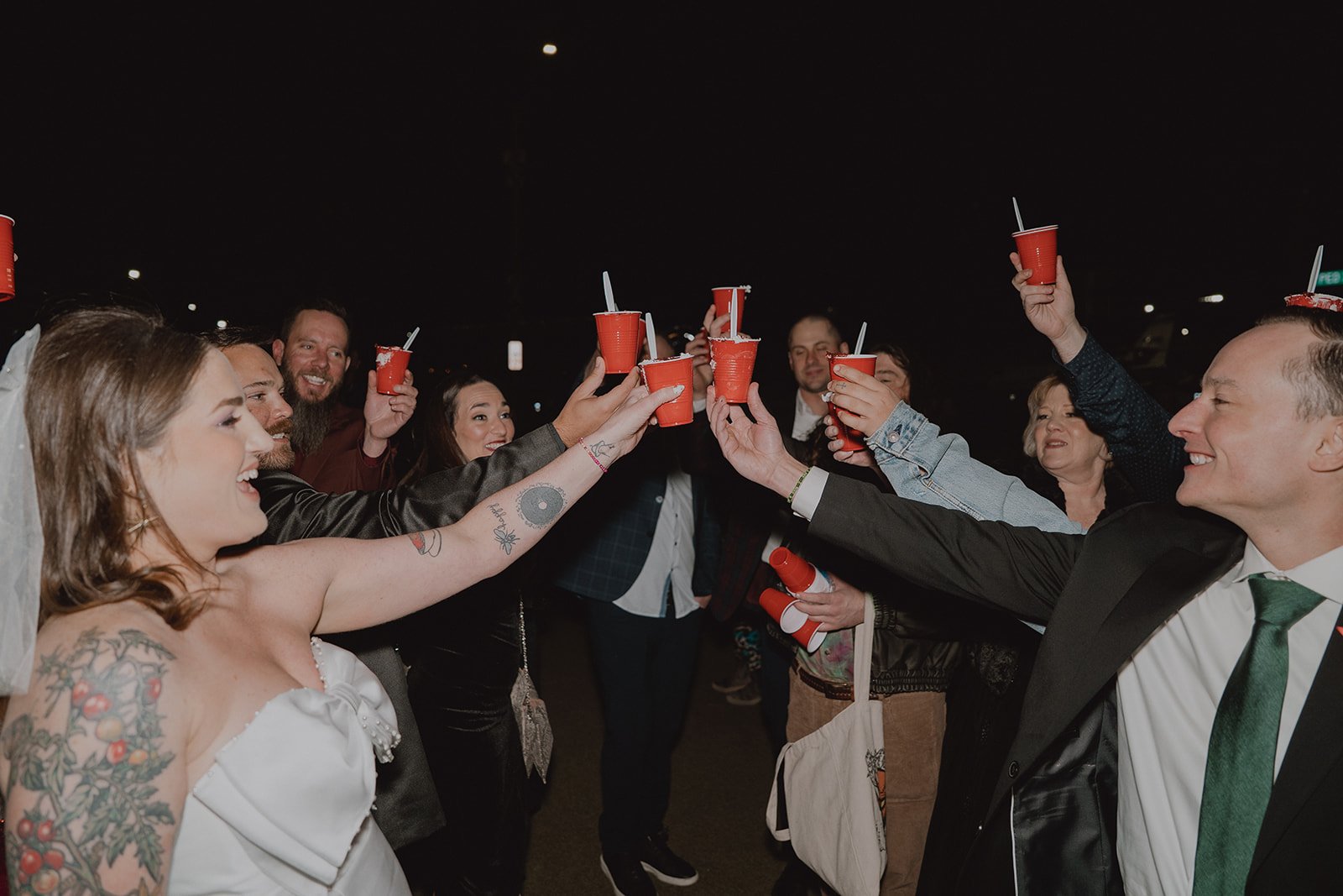 Couple clinking red solo cups with wedding guests in front of the Welcome to Fabulous Las Vegas sign at night.