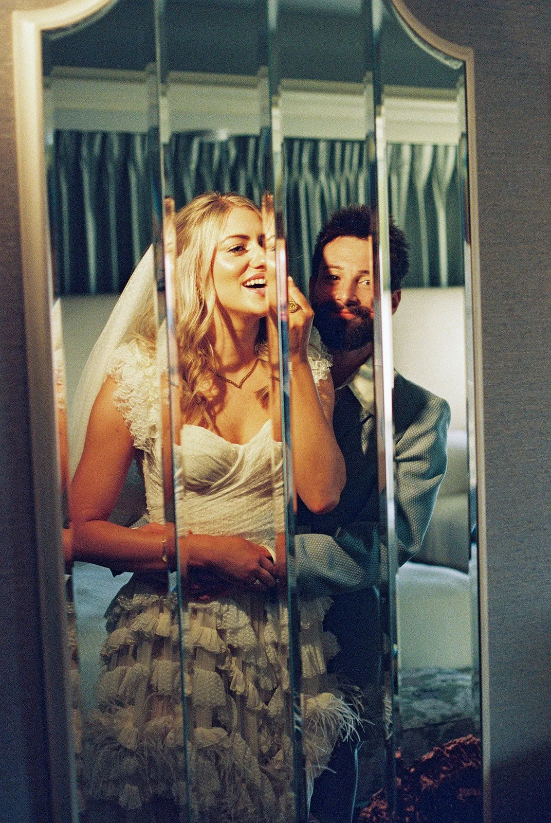 Bride applying lipstick on in the mirror with the groom at Couple having champagne during their elopement inside the Jackie Gaughan Suite at El Cortez Hotel in Las Vegas on film.
