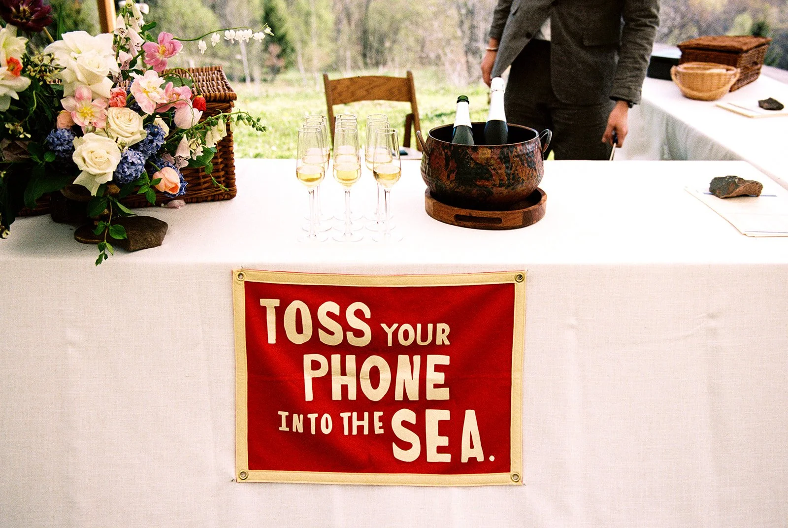 Champagne table and a sign that says "toss your phone into the sea" for an outdoor mountain wedding ceremony.