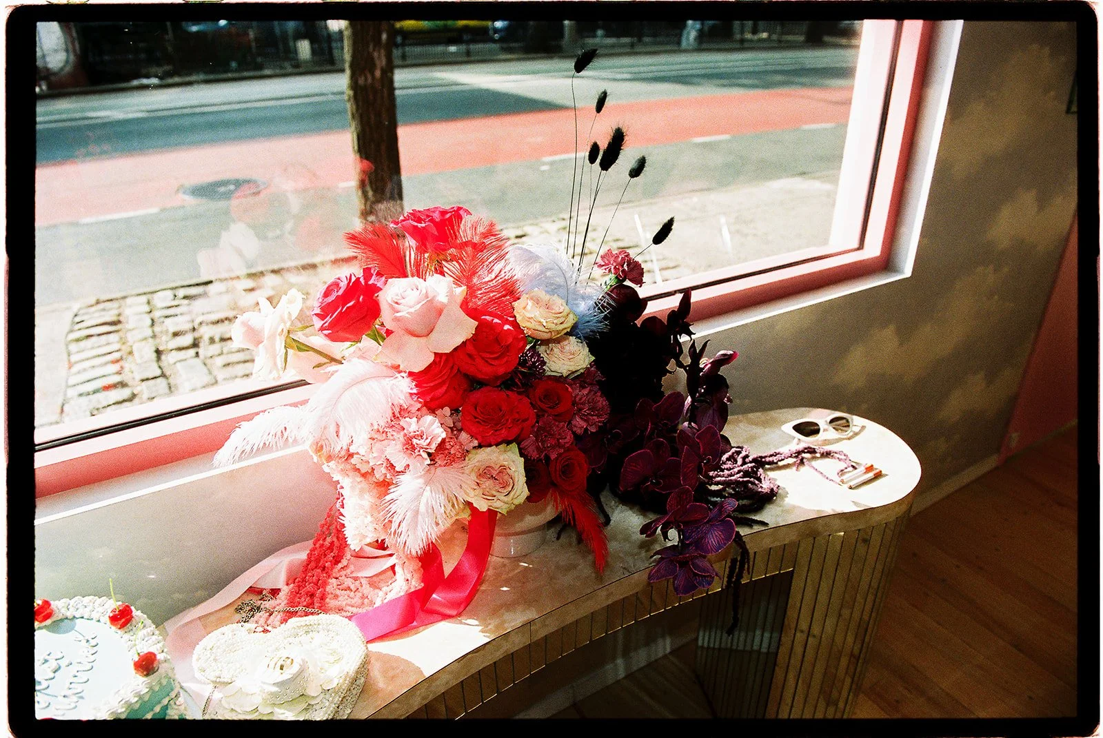 Colorful wedding flower arrangement with red and pink roses, dark orchids, and feathers beside a window at Sure Thing NYC.