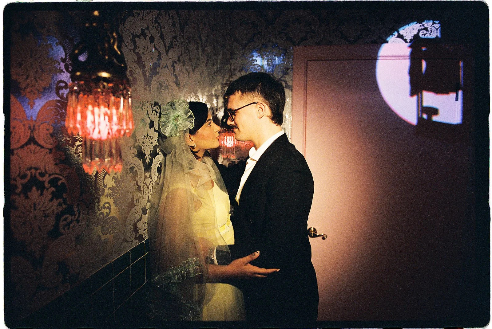 Couple standing face-to-face beside ornate vintage wallpaper and a glowing red wall sconce at Sure Thing NYC.