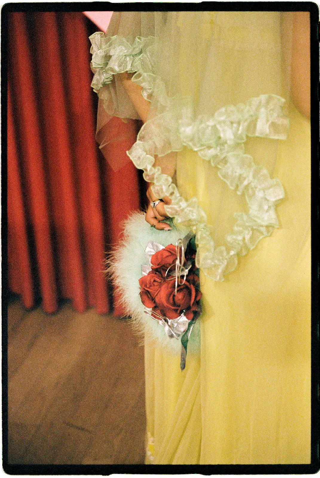Close-up of a pale yellow wedding dress with ruffled lace and a red rose bouquet against red velvet curtains.