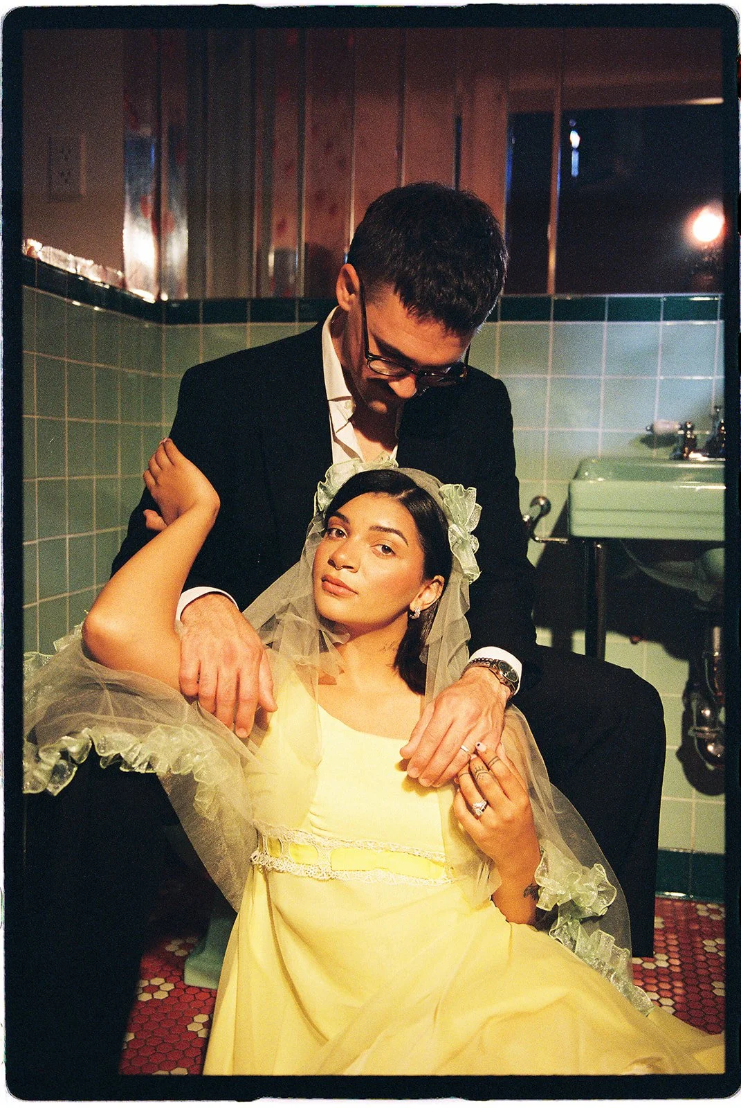 Couple posing together in a vintage tiled bathroom, with the bride wearing a yellow wedding dress and ruffled veil.