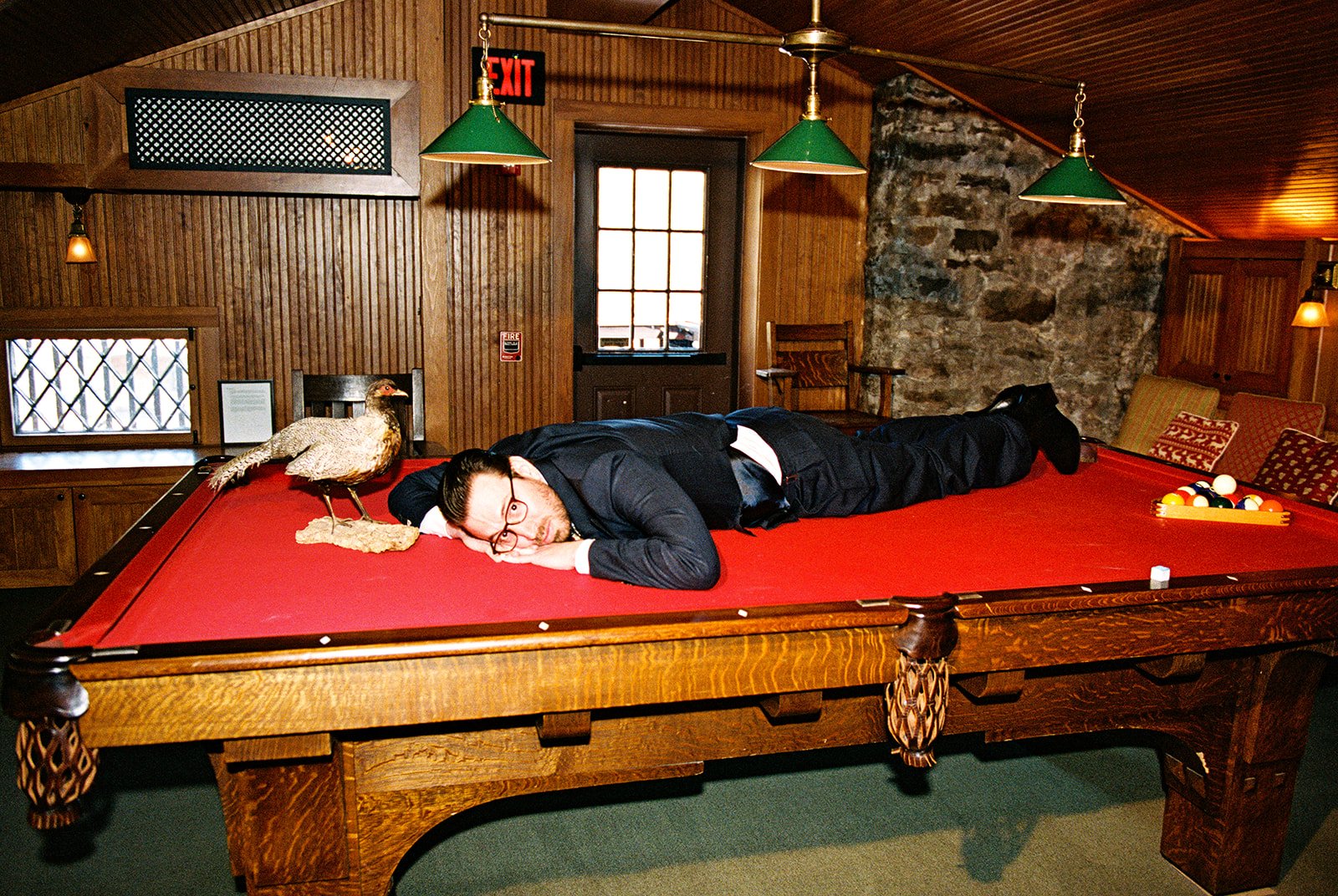 Playful film photo of the groom lying on the red pool table beside a taxidermy bird inside the Deer Mountain Inn Attic.