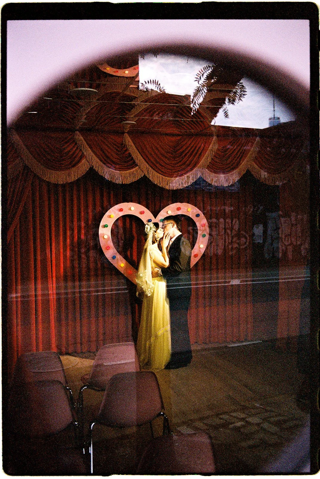 Couple kissing beneath a pink heart-shaped wedding arch, photographed through the window of Sure Thing NYC.
