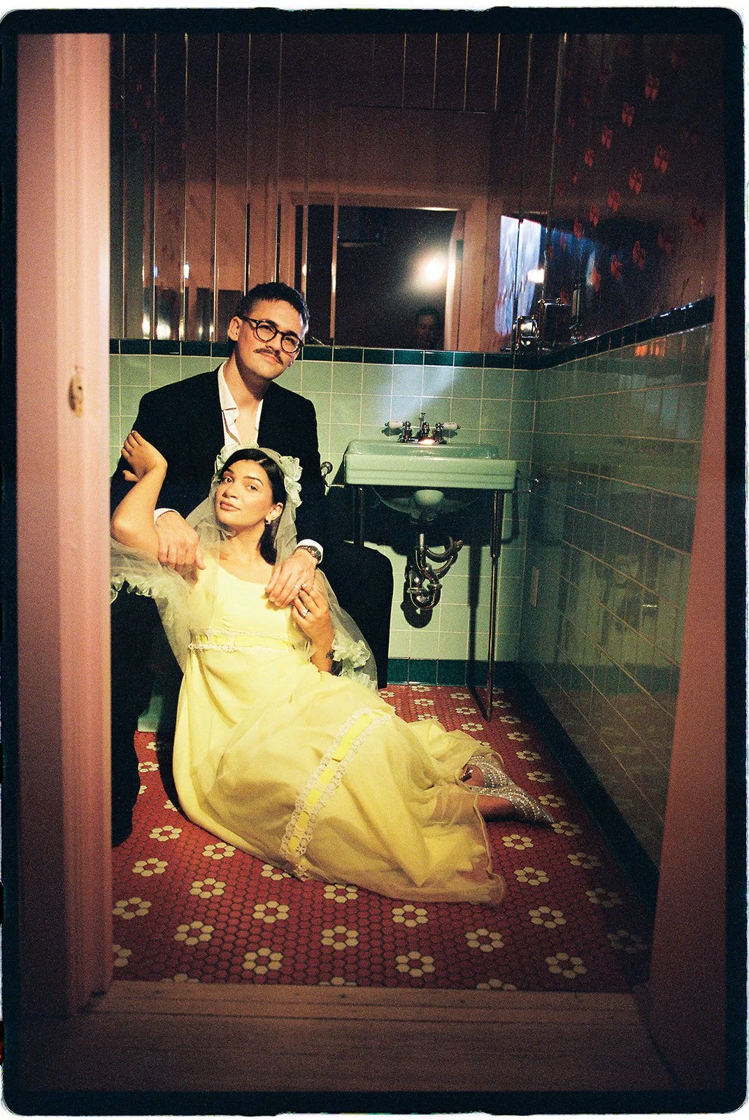 Couple posing in a retro bathroom with mint green tiles, a vintage sink, and red patterned flooring at Sure Thing NYC.