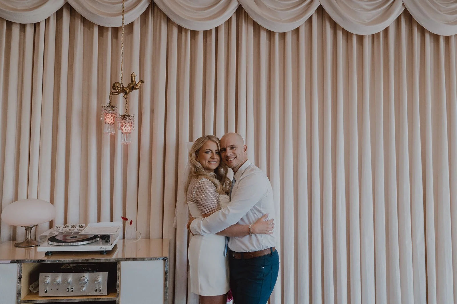 Newlyweds hugging inside Sure Thing Chapel with vintage curtains and record player behind them.