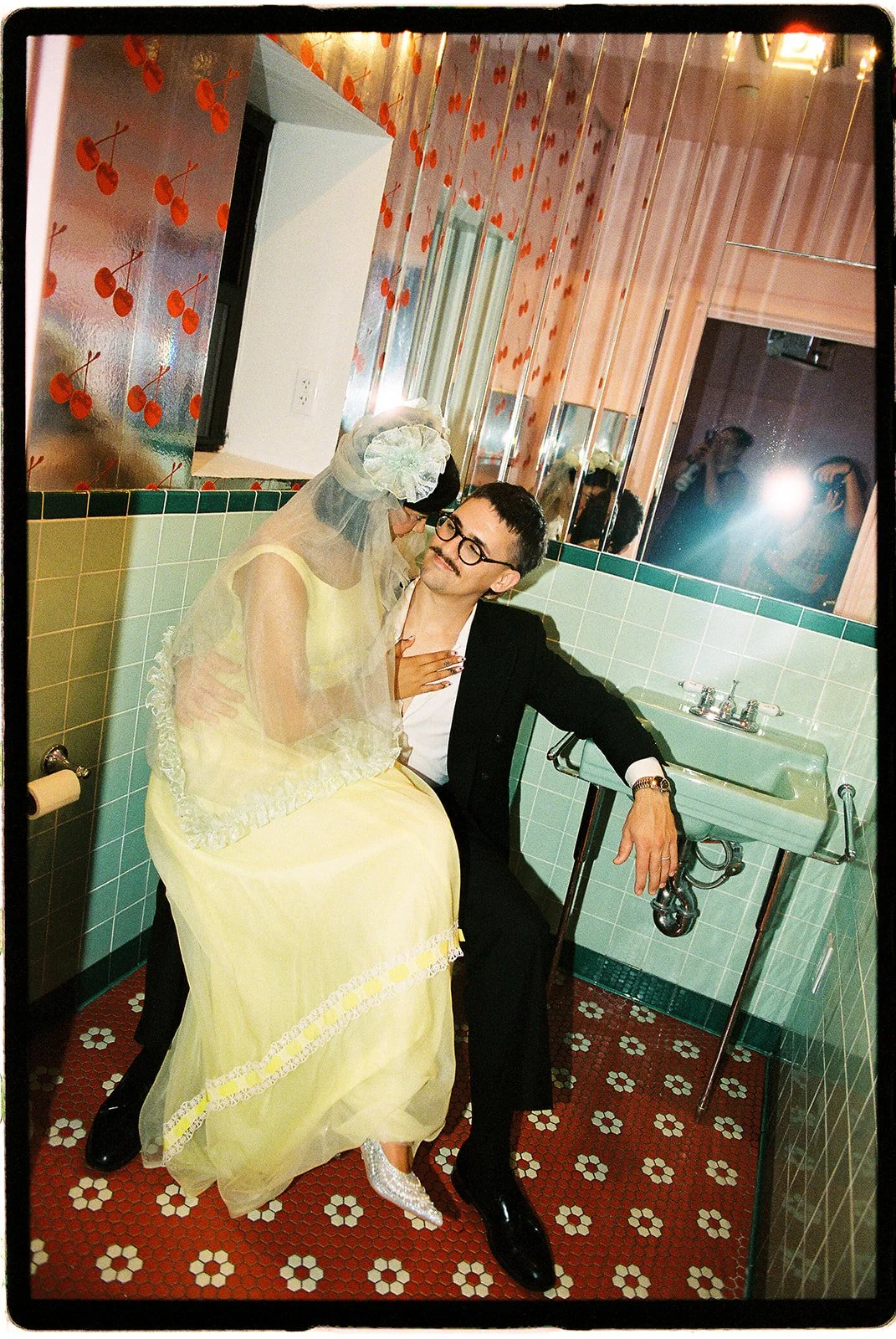 Couple sharing an intimate moment in a retro bathroom with cherry wallpaper, mint green tiles, and a vintage sink.
