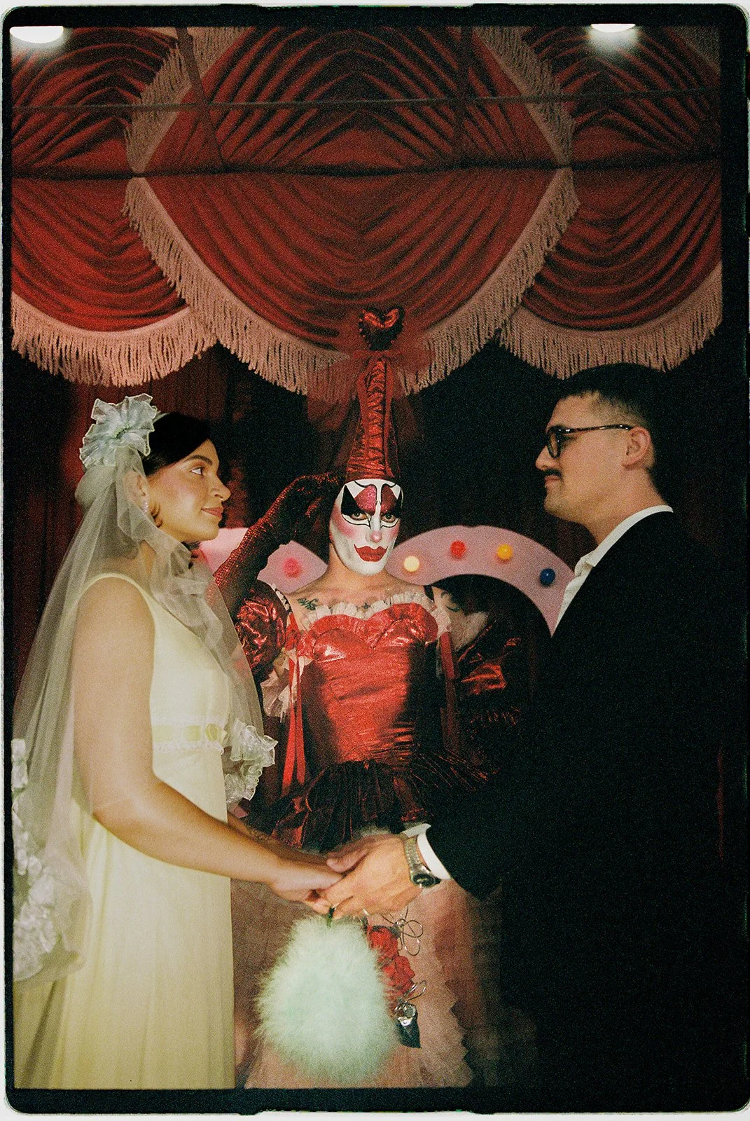 Intimate wedding ceremony at Sure Thing NYC with Tillie the Clown standing between the couple beneath red velvet drapery.
