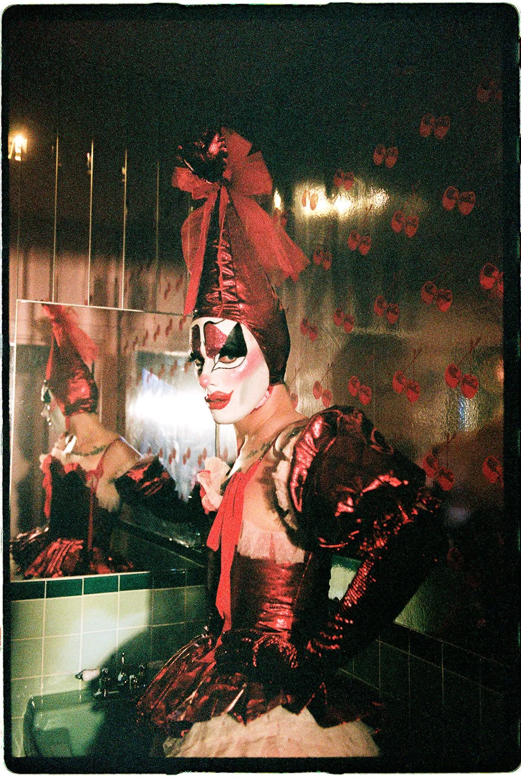 Tillie the Clown posing in a red theatrical costume and tall pointed hat beside a mirror at Sure Thing NYC.