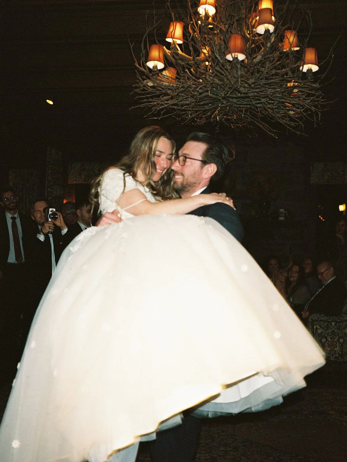 Playful film photo of the groom carrying the bride beneath the branch chandelier during their Deer Mountain Inn reception.