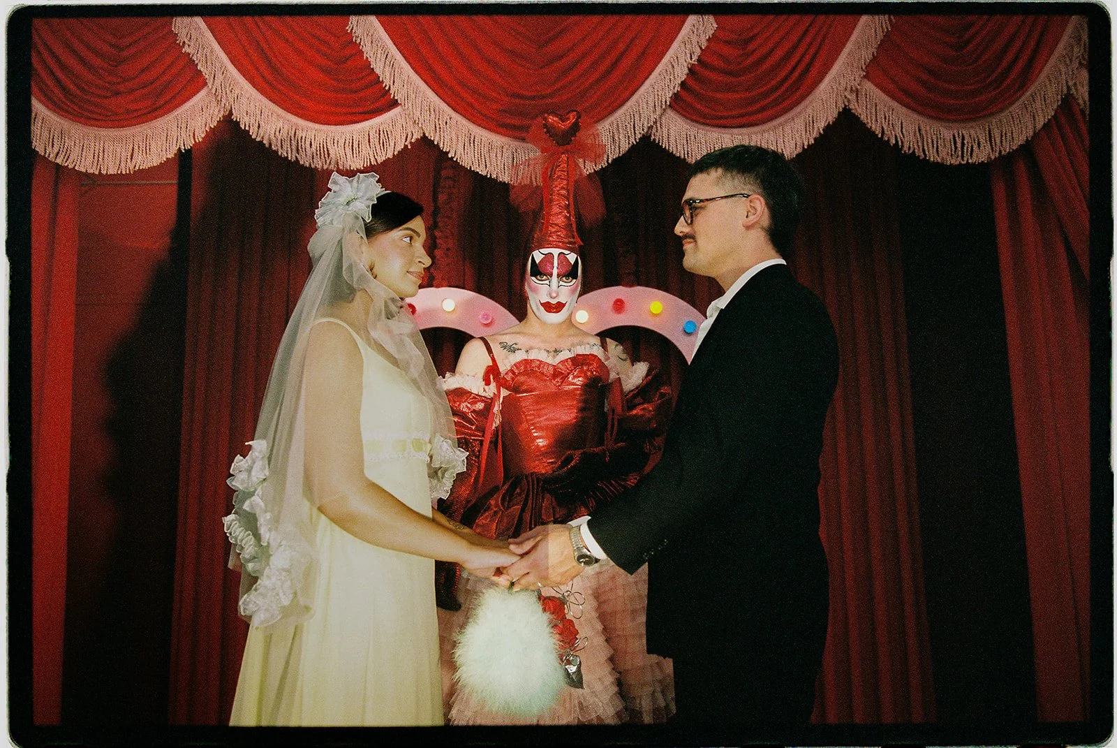 Couple holding hands during their Sure Thing NYC wedding ceremony with Tillie the Clown officiating in front of red curtains.