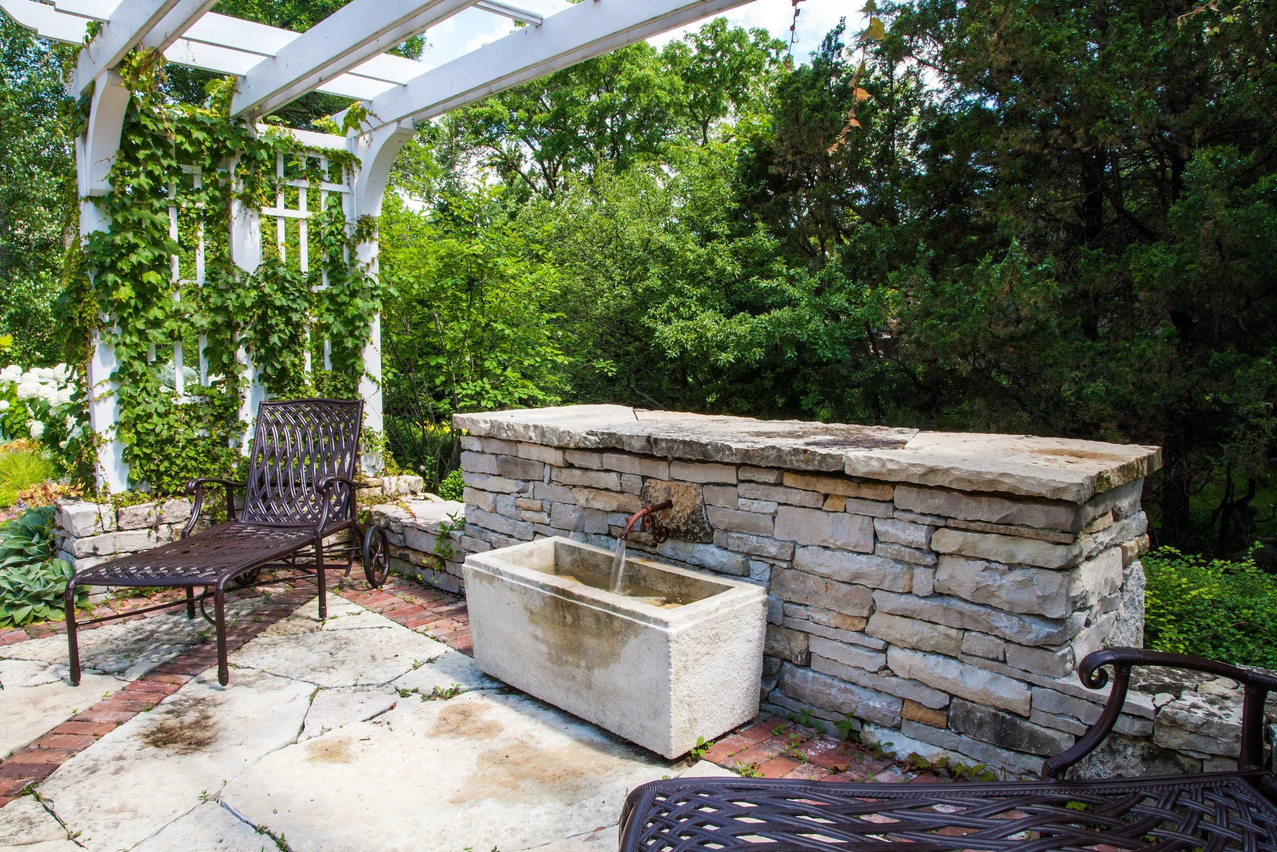 A garden patio with stone flooring, metal chairs, a built-in stone water fountain, and a white wooden pergola covered in green ivy, surrounded by lush green trees.