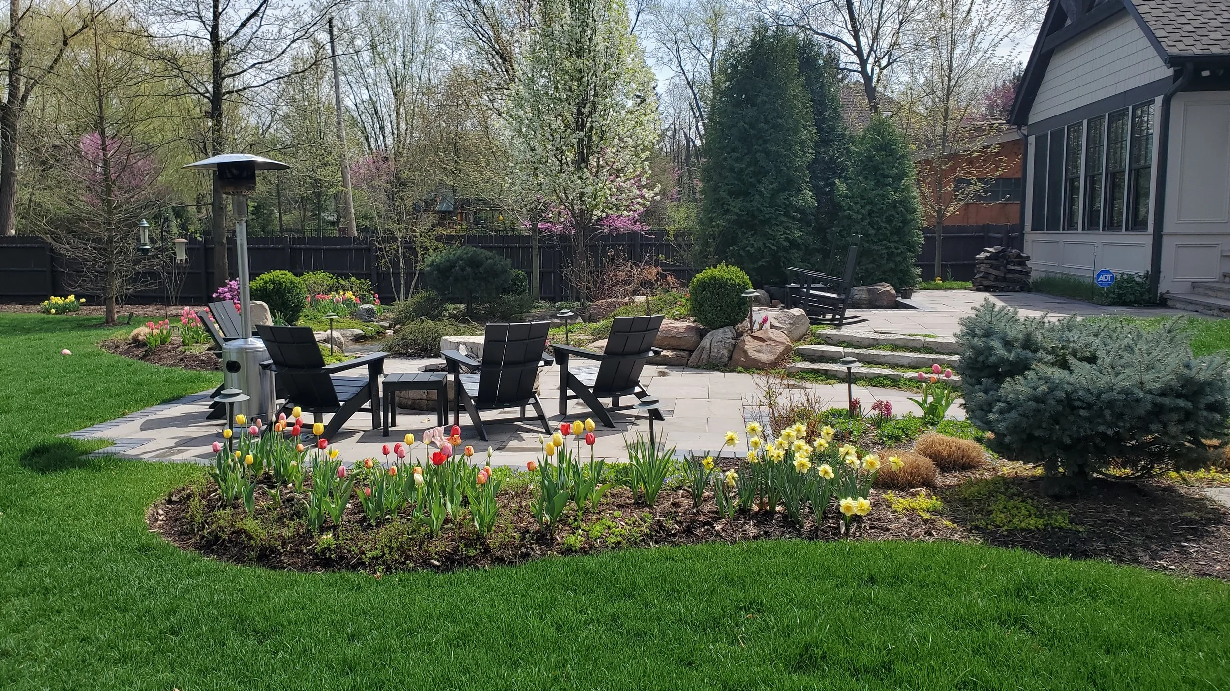 A landscaped backyard with a patio featuring outdoor chairs, a heater, and flower beds with blooming tulips and daffodils, surrounded by trees and a fence.