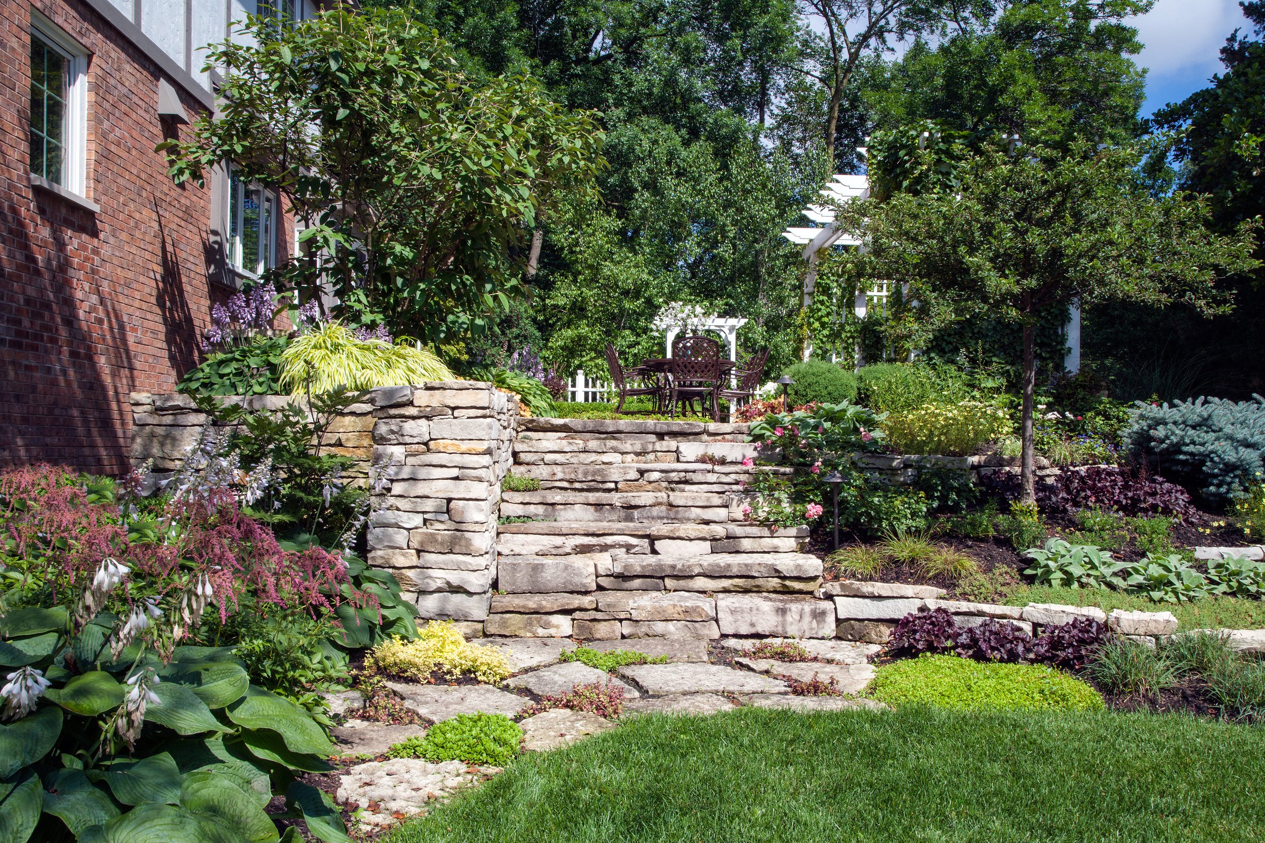 A landscaped backyard garden with stone steps leading up to a patio with a table and chairs, surrounded by lush greenery, trees, and various flowering plants.