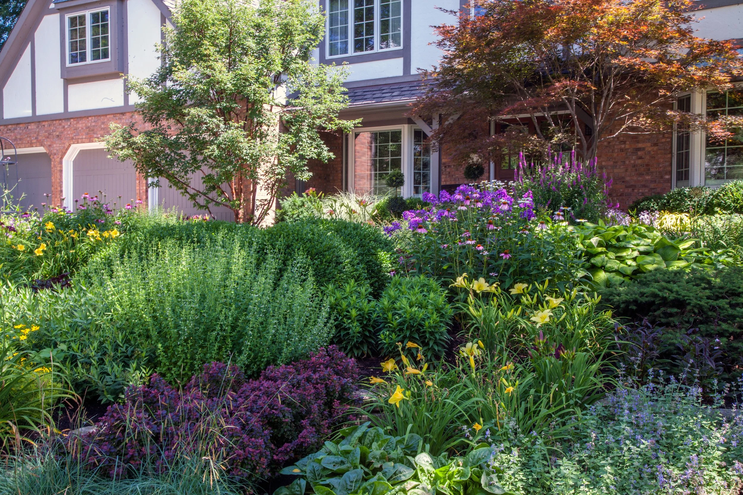 Front yard garden with a variety of colorful flowers and green shrubs in front of a house with large windows and brick walls.