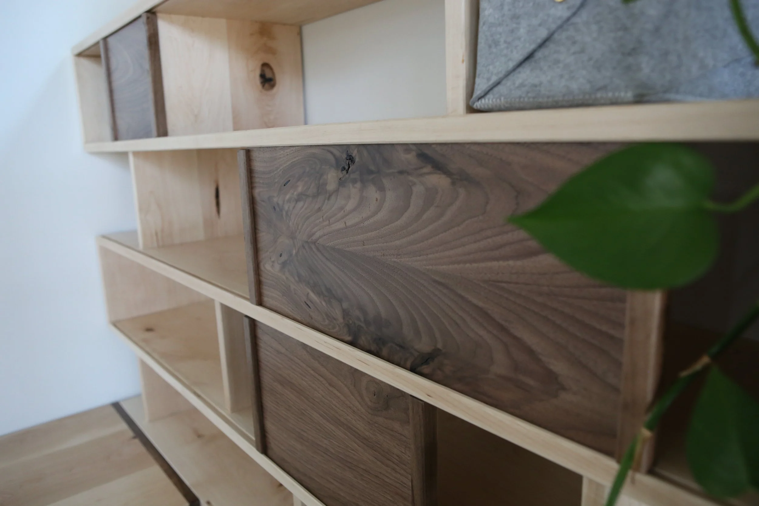Wooden shelf with compartments in various shades of brown and natural wood tone, with a gray cloth or cushion and some green plant leaves visible in the corner.