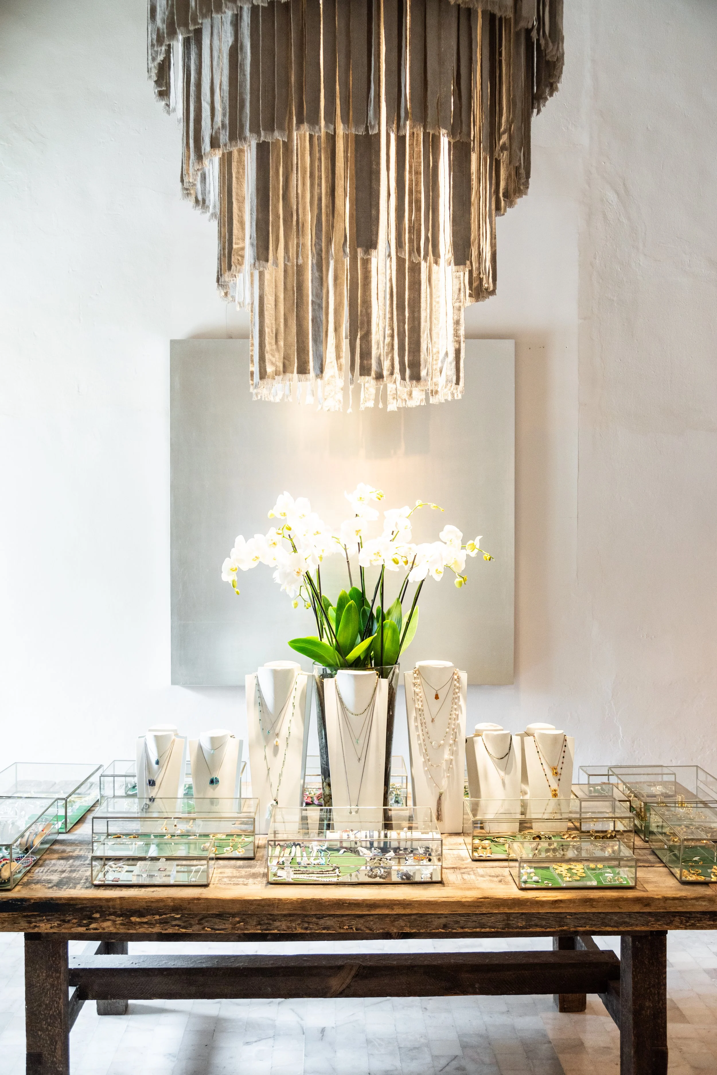 Mesa de madera con exhibición de joyas y collares, con un jarrón grande con orquídeas blancas en el centro y un araña de tela suspendida del techo.
