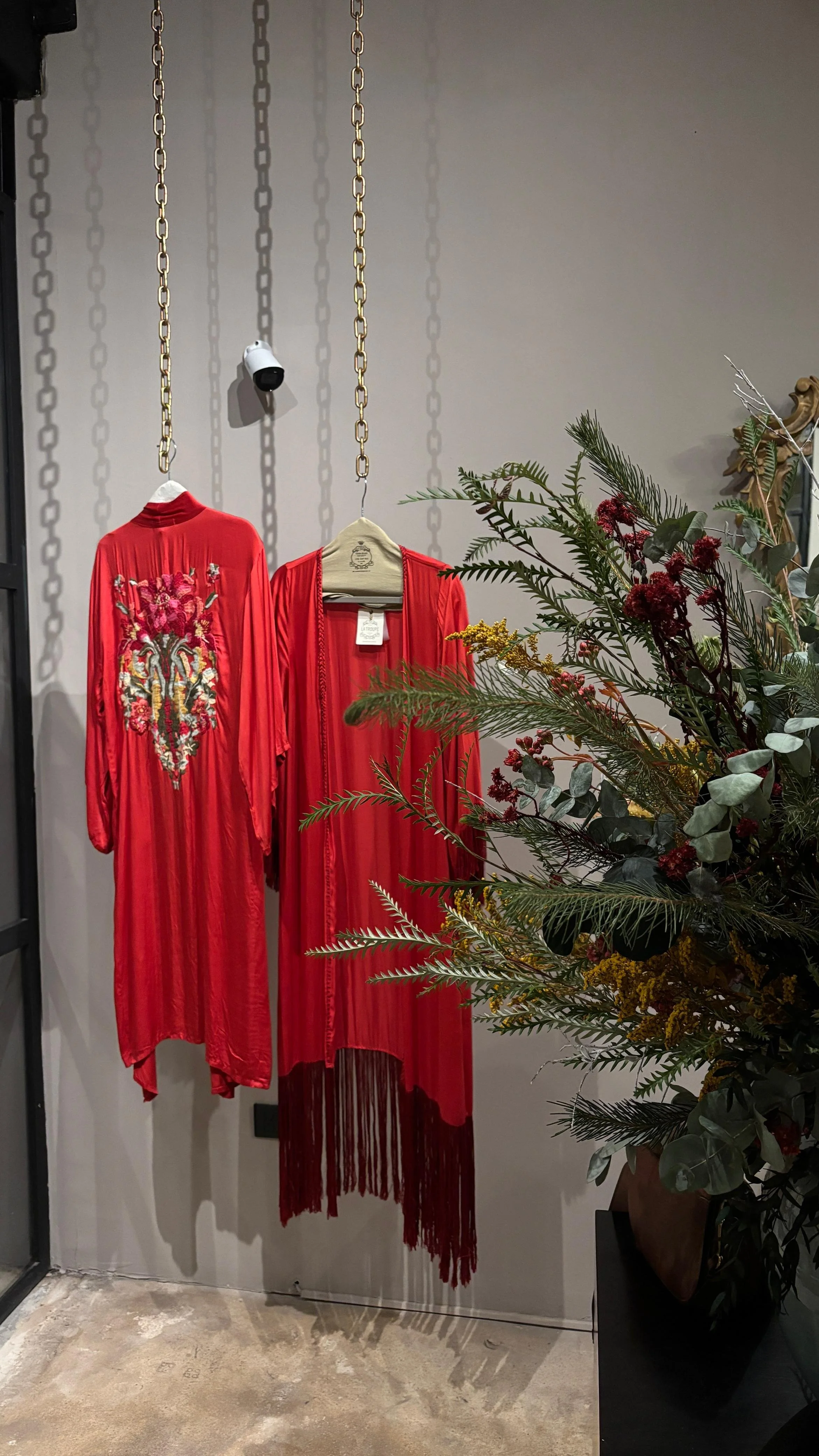 Vestidos largos rojos con detalles de encaje y bordado en exhibición, decorados con cadenas doradas en un fondo gris, junto a un arreglo floral con diferentes tipos de follaje y flores.