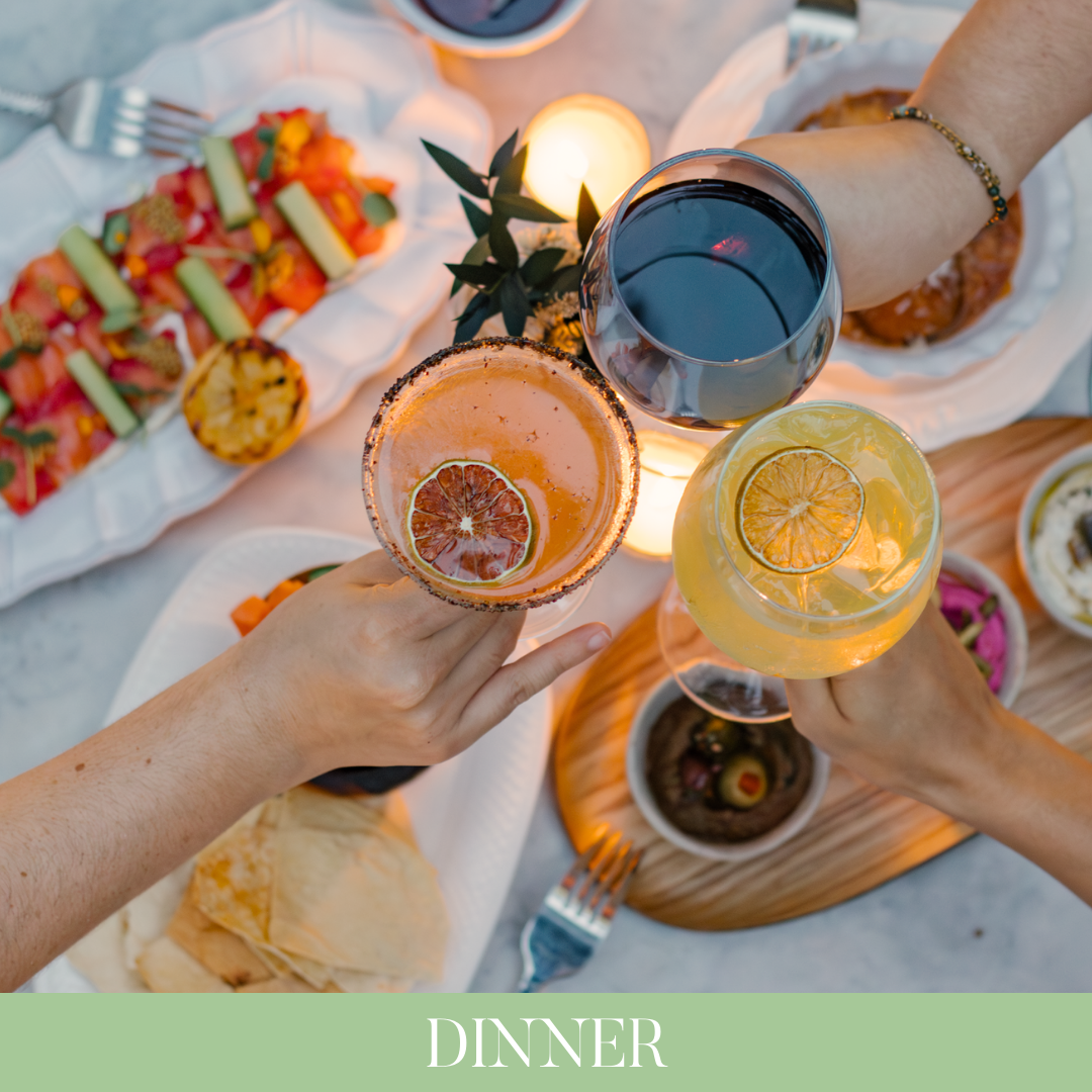 Personas brindando con bebidas en una mesa con comida. Hay platos con comida, copas con vino y cócteles, y una vela encendida en la mesa.