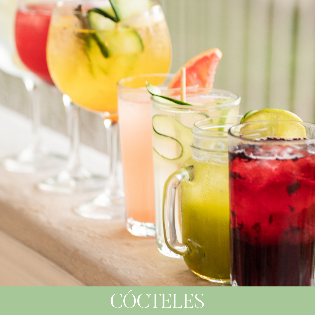 Una variedad de cócteles coloridos con frutas frescas y rodajas de limón, pepino, sandía y agua de jamaica en vasos y jarros en una mesa.