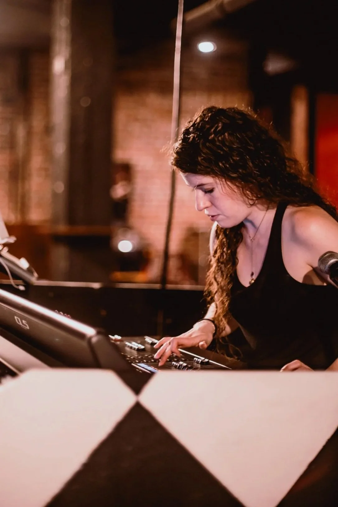 A woman with curly brown hair operating a digital mixing console in a dimly lit, brick-walled venue.