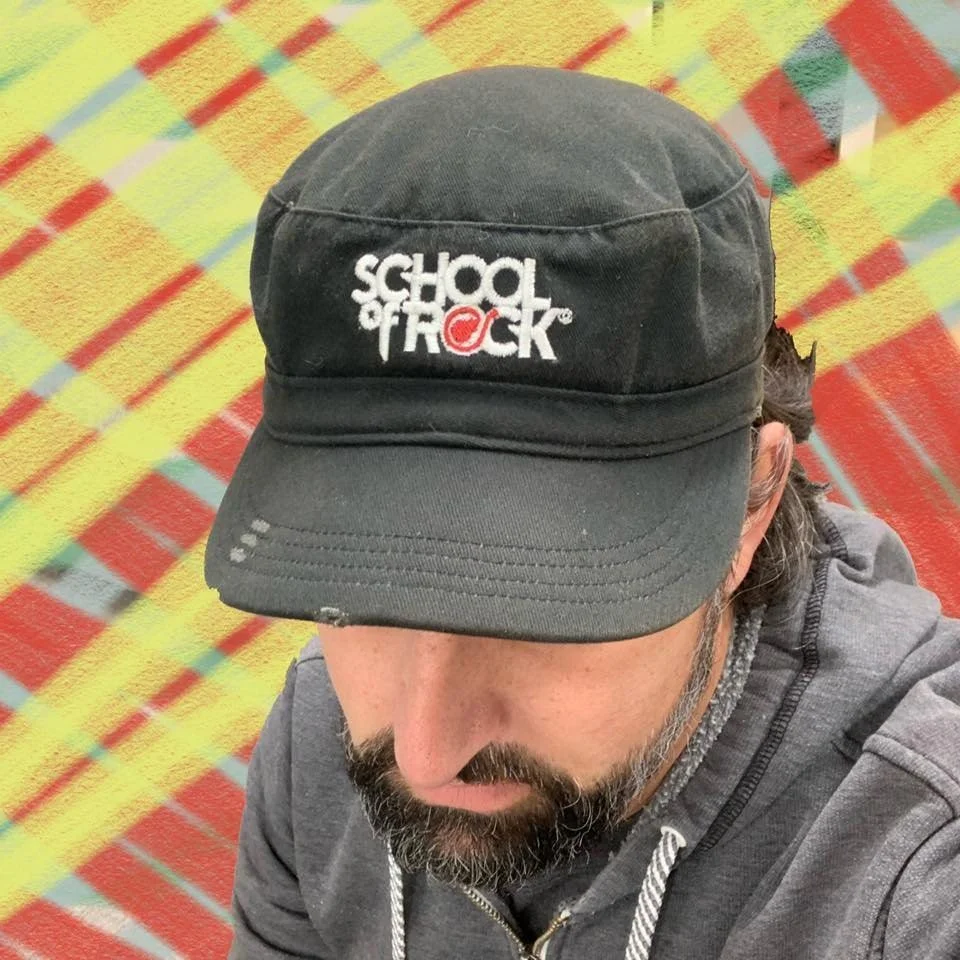 A man with a beard wearing a black cap with white and red text saying 'School of Rock' on a colorful, geometric patterned floor.