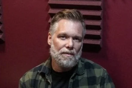 A man with a beard and mustache wearing a green plaid shirt, sitting in front of a maroon wall with acoustic foam panels.