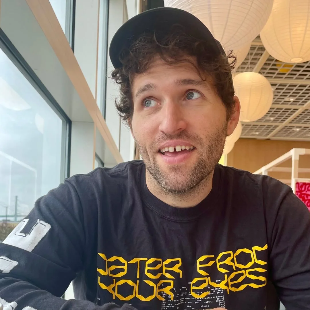 A man with curly hair, beard, and mustache wearing a black cap and black T-shirt, sitting indoors near large windows and a ceiling decorated with paper lanterns.