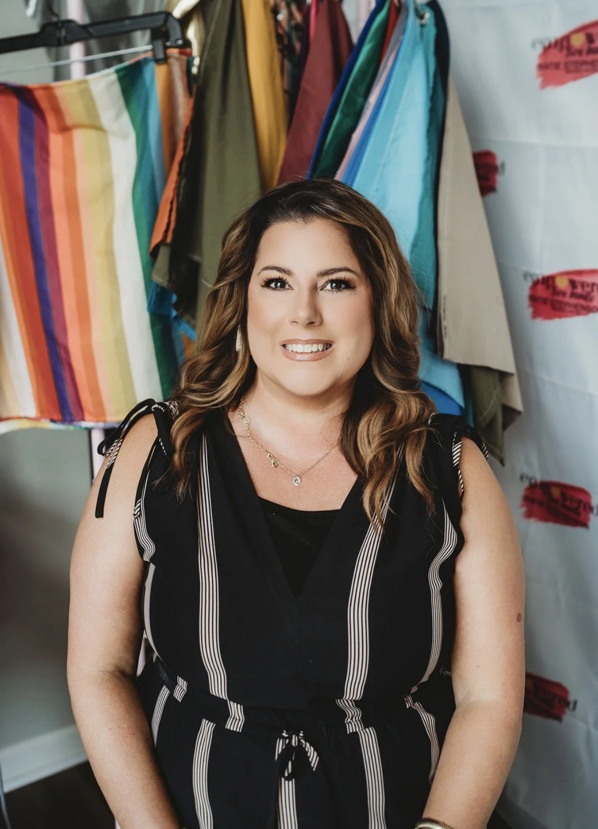 A woman with wavy brown hair, smiling, wearing a black and white striped dress, standing in front of colorful fabric swatches in a store or workshop.