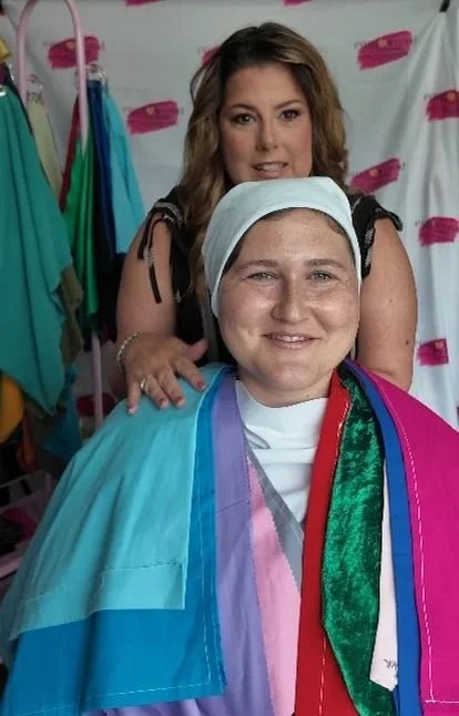 A woman with short hair and a broad smile, dressed in a colorful gown and head covering, sitting in a salon chair. Behind her is another woman with long brown hair, smiling and posing. The background shows a salon setting with colorful aprons hanging.