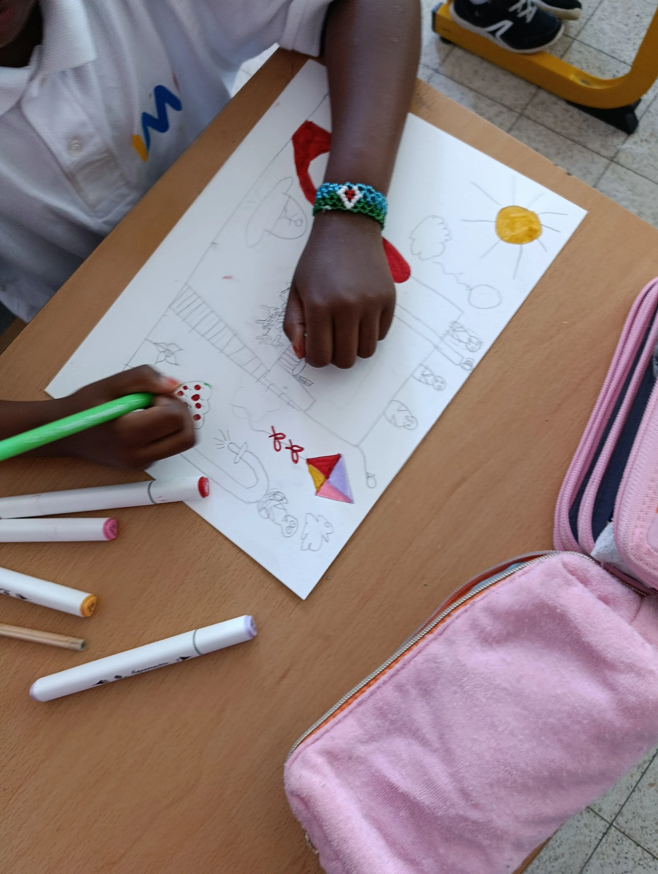 Une main d'enfant en train de colorier une feuille blanche avec des dessins d'arc-en-ciel, de soleil, de maison et d'animaux, sur un bureau en bois. Plusieurs marqueurs colorés sont posés autour de la feuille. La personne porte un bracelet en perles multicolores, et un sac à main rose est également visible.