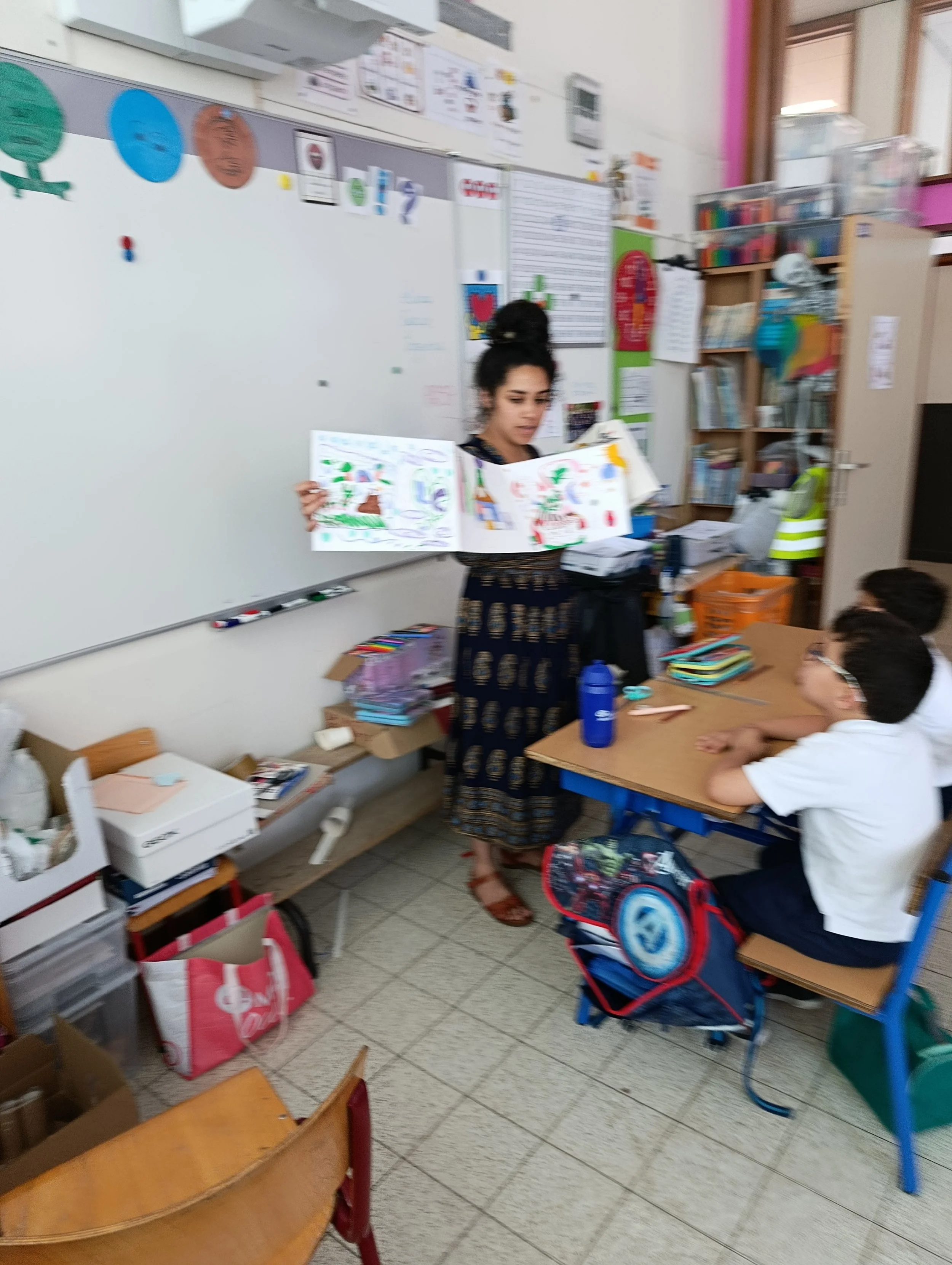 Une jeune fille debout devant un tableau blanc en classe, tenant un livre coloré ouvert, avec deux garçons assis à un bureau, un sac à dos à leurs côtés, dans une salle de classe avec des étagères contenant des livres, des affiches et un tableau d'affichage.