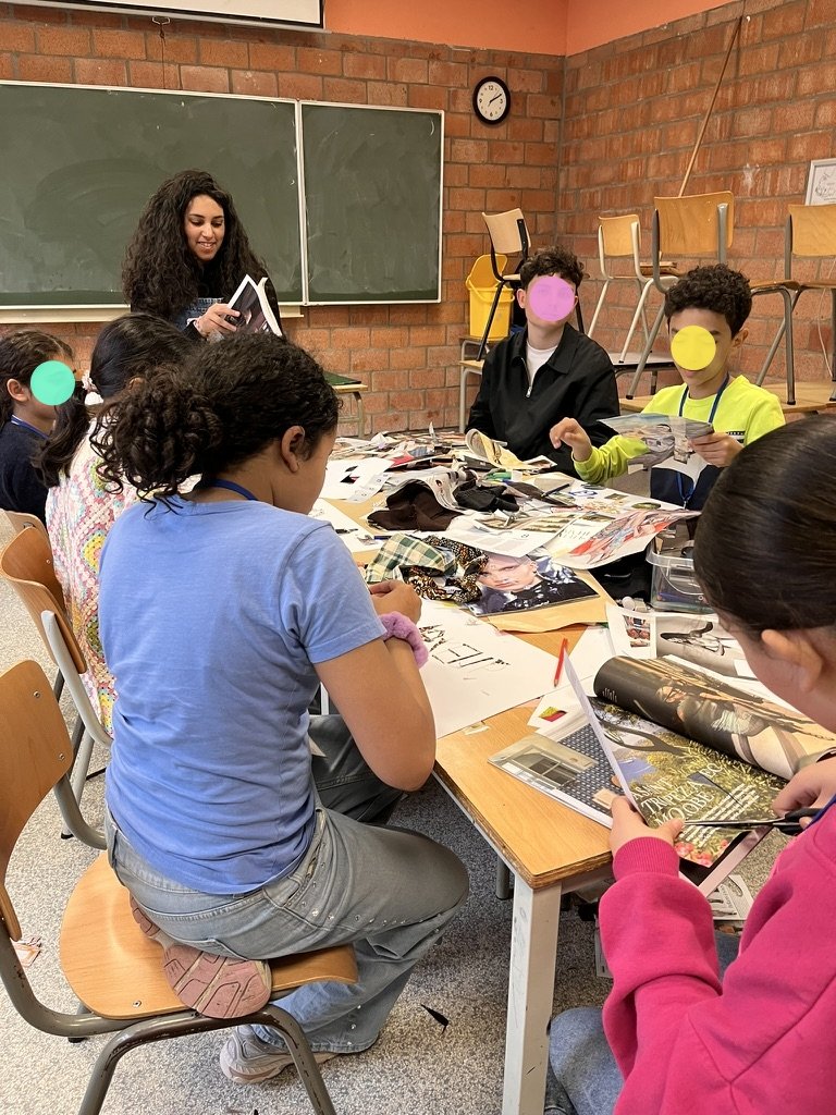 Groupe d'élèves en classe, certains lisant des magazines ou des livres, une femme semble leur donner une explication ou présenter quelque chose, dans une salle avec un tableau vert et un mur en briques.
